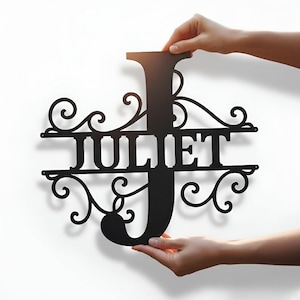 Puede incluir: Un letrero de metal negro con el nombre "JULIET" en una fuente decorativa. La letra "J" es grande y estilizada, con adornos ornamentados arriba y abajo del nombre. El letrero se sostiene sobre un fondo blanco.