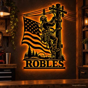 May include: A black metal wall sign with warm orange backlighting featuring a silhouette of a utility worker on a power pole, an American flag, and the name ROBLES in bold capital letters at the bottom of the rectangular design.