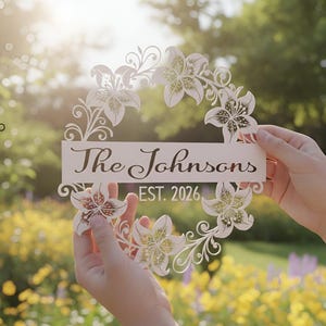 Puede incluir: Una corona floral blanca cortada con l&aacute;ser con el texto "The Johnsons EST. 2026". La corona presenta dise&ntilde;os detallados de flores de lirio y se sostiene frente a un fondo borroso al aire libre.