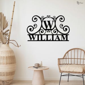 Puede incluir: Arte mural de metal negro con el nombre "WILLIAM" y un diseño ornamental. La letra "W" está centrada en un círculo. La obra está montada en una pared blanca, con un jarrón y una mesa auxiliar de tonos neutros en primer plano.