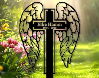 May include: A black metal memorial cross with angel wings. The cross features the name "Ellie Hamm" and the years "1954-2025". The wings are intricately cut out, creating a delicate design. The memorial is set in a garden.
