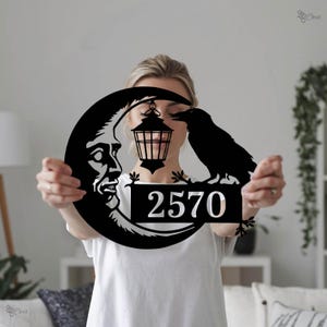 Pode incluir: Placa de endereço em metal preto com lua crescente com rosto, lanterna e corvo. O número 2570 é exibido em branco. Design decorativo para uso externo.