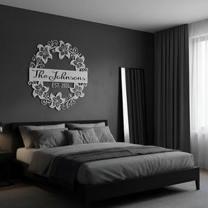 Puede incluir: Una corona floral de metal blanco con el texto "The Johnsons EST. 2026" montada en una pared gris oscuro sobre una cama. La corona presenta un dise&ntilde;o circular con detalles de flores y hojas.