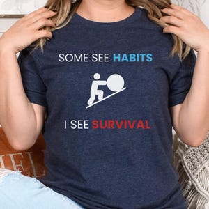 May include: A navy blue t-shirt with a white graphic of a person pushing a ball up a hill. The text "Some See Habits I See Survival" is printed on the shirt.
