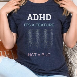 Peut inclure: Un t-shirt bleu marine avec le texte "ADHD It's a feature Not a bug" en blanc. Le t-shirt présente un motif de grille de code binaire en bleu clair.