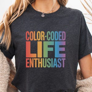 Peut inclure: T-shirt gris foncé avec l'inscription "COLOR-CODED LIFE ENTHUSIAST" en dégradé arc-en-ciel. Le t-shirt à manches courtes semble être fait d'une matière douce. Le texte est en gras, sans empattement.