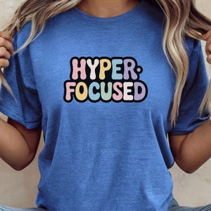 May include: Blue short-sleeved t-shirt with the text "HYPER FOCUSED" in a colourful, retro font. The text is outlined in black and features pink, yellow, blue, and green letters. The shirt is made of a soft material.