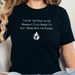 May include: Black t-shirt with white text that reads "I've Hit the Point in the Burnout Cycle Where I'm Just Vibing With the Flames." A white flame graphic is below the text. The shirt is a crew neck style.