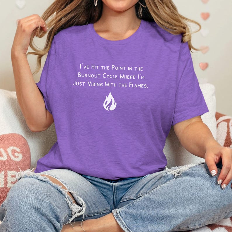 May include: A purple t-shirt with the text "I'VE HIT THE POINT IN THE BURNOUT CYCLE WHERE I'M JUST VIBING WITH THE FLAMES." and a flame graphic. The shirt is paired with blue jeans.