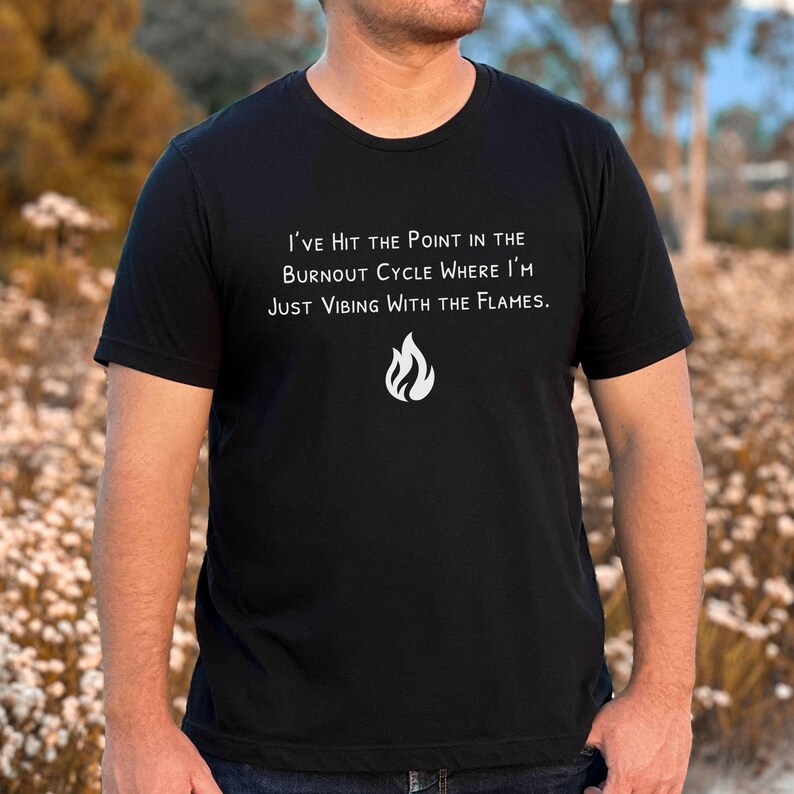 May include: Black t-shirt with white text that reads "I'VE HIT THE POINT IN THE BURNOUT CYCLE WHERE I'M JUST VIBING WITH THE FLAMES." A white flame graphic is below the text. The shirt is a crew neck.