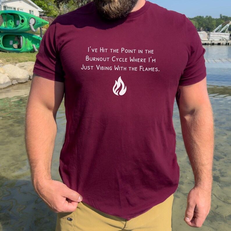 May include: Maroon t-shirt with white text: "I'VE HIT THE POINT IN THE BURNOUT CYCLE WHERE I'M JUST VIBING WITH THE FLAMES." A white flame graphic is below the text. The shirt is short-sleeved.
