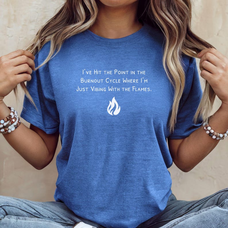 May include: Blue t-shirt with the text "I'VE HIT THE POINT IN THE BURNOUT CYCLE WHERE I'M JUST VIBING WITH THE FLAMES." and a white flame graphic. The shirt is a crew neck style and has short sleeves.