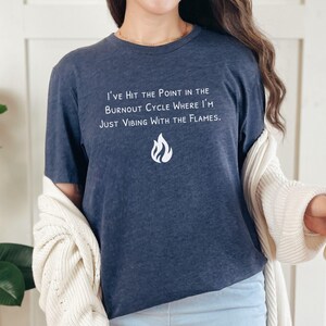 May include: Navy blue t-shirt with white text: "I've Hit the Point in the Burnout Cycle Where I'm Just Vibing With the Flames." A flame graphic is below the text. The shirt is a casual wear item.