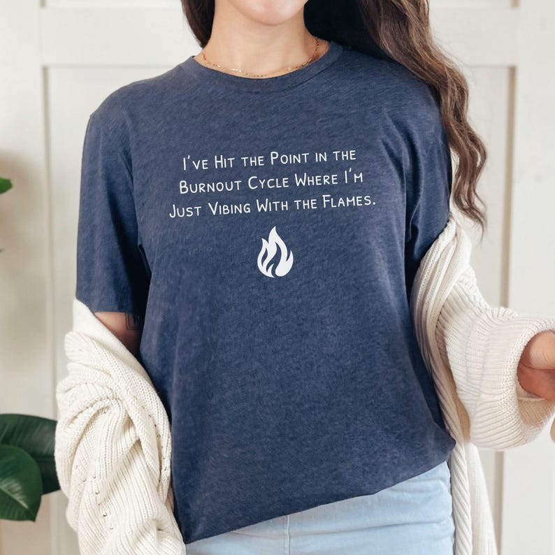 May include: Navy blue t-shirt with white text: "I've Hit the Point in the Burnout Cycle Where I'm Just Vibing With the Flames." A flame graphic is below the text. The shirt is a casual wear item.