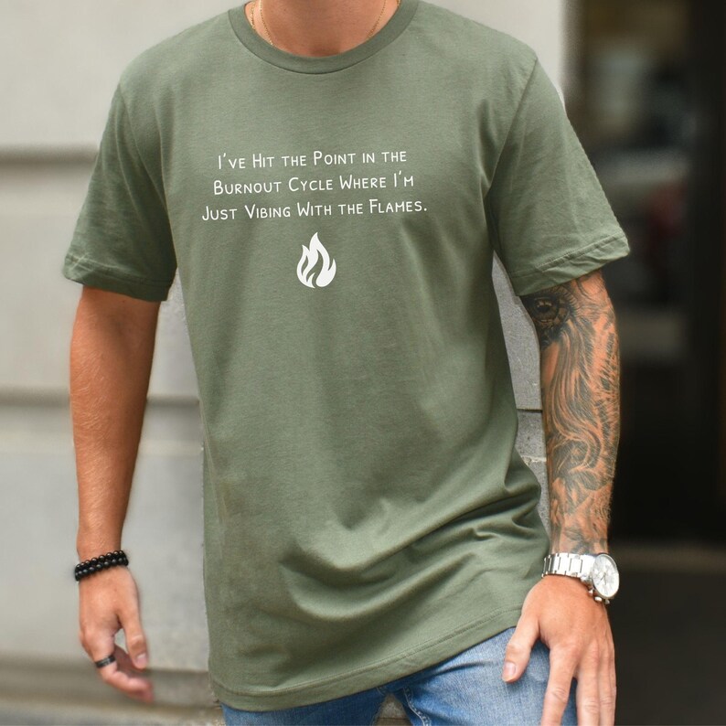 May include: Olive green t-shirt with white text: "I'VE HIT THE POINT IN THE BURNOUT CYCLE WHERE I'M JUST VIBING WITH THE FLAMES." A white flame graphic is below the text. The shirt is short-sleeved.