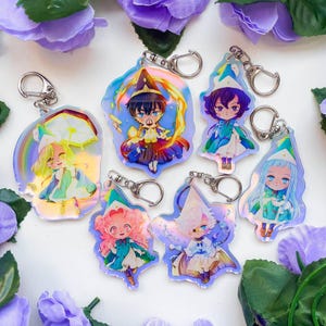 Könnte beinhalten: Sechs schimmernde Schlüsselanhänger mit Anime-Charakteren in bunten Outfits und fantasievollen Hüten. Jeder Schlüsselanhänger hat einen silbernen Ring und eine holografische Oberfläche.