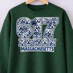 May include: A dark green sweatshirt with the number "617" in white and blue, filled with a pattern of soccer balls. Below the number, the word "BOSTON" is displayed, and below that, "MASSACHUSETTS" in white letters.