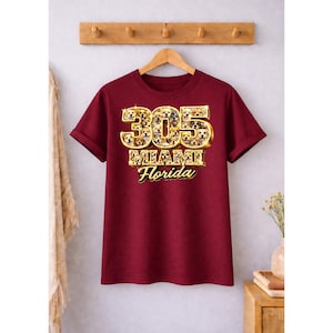 Puede incluir: Una camiseta granate con las mangas enrolladas. La parte delantera presenta el texto "305 Miami Florida" en una fuente dorada y texturizada. Los números están rellenos con un patrón de pequeñas caras de perro.