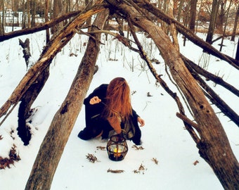 May include: A person kneeling in the snow, holding a lit lantern. The scene is framed by a structure of wooden branches, with a snowy forest background. The lantern emits a warm glow.