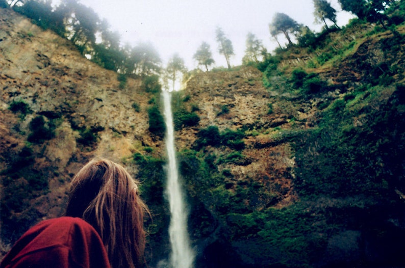 May include: A person with long brown hair stands in front of a tall waterfall cascading down a rocky cliffside. Lush green foliage covers the cliff face, creating a natural backdrop for the scene.