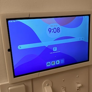 May include: A white tablet with a blue and purple background is mounted on a wall. The tablet's screen displays the time, "9:08", and the date, "Wed, Jan 12". The screen also shows a search bar with the Google logo and a Facebook icon. There are four app icons at the bottom of the screen.