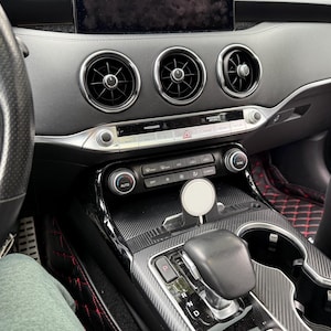 May include: Interior view of a car dashboard. The dashboard is black with silver accents, featuring air vents, a central console with climate controls, and a gear shift. A steering wheel is visible on the left. A wireless charger is in the center console.