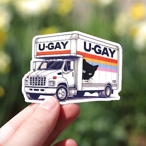 Puede incluir: Una pegatina blanca de un cami&oacute;n de mudanzas con el texto "U-GAY" en la parte superior y un gato negro asom&aacute;ndose. El cami&oacute;n tiene un dise&ntilde;o de rayas arco&iacute;ris. La pegatina se sostiene frente a un fondo borroso de flores amarillas.