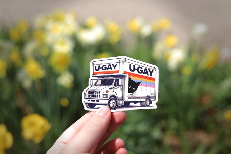 Puede incluir: Una pegatina blanca de un cami&oacute;n de mudanzas con el texto "U-GAY" en la parte superior y un gato negro asom&aacute;ndose. El cami&oacute;n tiene un dise&ntilde;o de rayas arco&iacute;ris. La pegatina se sostiene frente a un fondo borroso de flores amarillas.