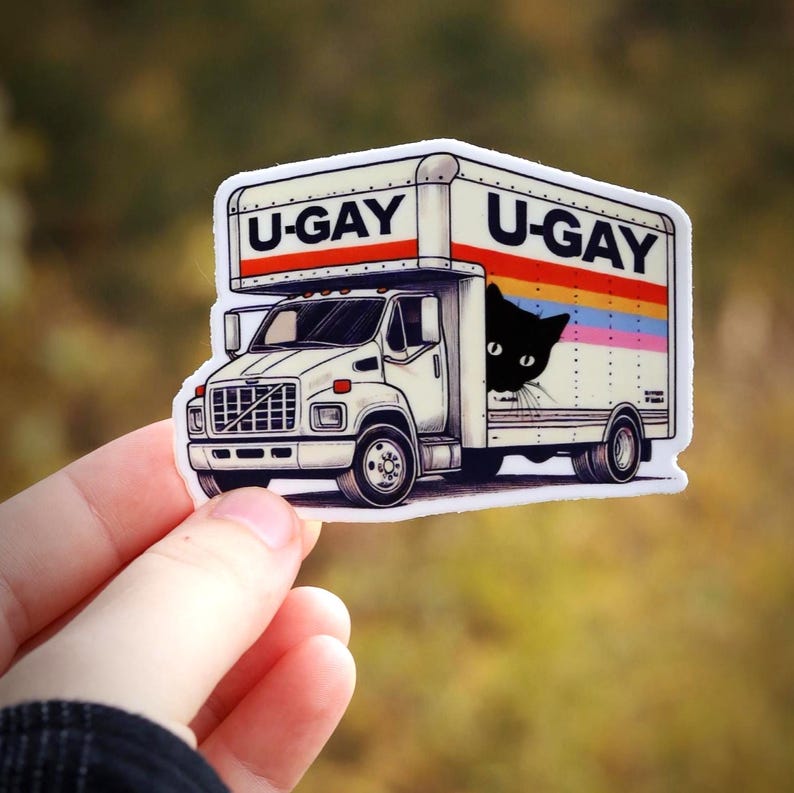 Puede incluir: Una pegatina que representa un cami&oacute;n de mudanzas blanco con el texto "U-GAY" en la parte superior y lateral. Un gato negro asoma por el lateral del cami&oacute;n, con un dise&ntilde;o de arco&iacute;ris. La pegatina est&aacute; sujeta por una mano.