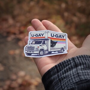 Puede incluir: Una pegatina blanca con un cami&oacute;n de mudanzas de dibujos animados con el texto "U-GAY" en la parte superior y lateral. Un gato negro asoma por el lateral del cami&oacute;n. El cami&oacute;n tiene un dise&ntilde;o de rayas arco&iacute;ris.