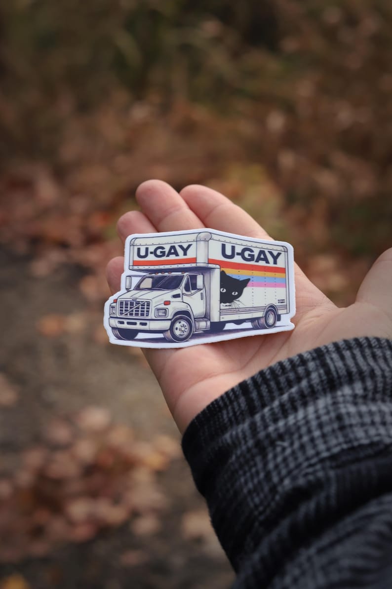 Puede incluir: Una pegatina blanca con un cami&oacute;n de mudanzas de dibujos animados con el texto "U-GAY" en la parte superior y lateral. Un gato negro asoma por el lateral del cami&oacute;n. El cami&oacute;n tiene un dise&ntilde;o de rayas arco&iacute;ris.