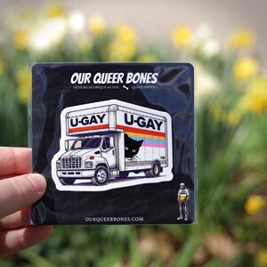Puede incluir: Adhesivo negro con un dise&ntilde;o de cami&oacute;n blanco con el texto "U-GAY" en la parte superior y los lados. Un gato negro asoma por la ventana del cami&oacute;n. El adhesivo tambi&eacute;n incluye el texto "OUR QUEER BONES" y "DESIGNS AS UNIQUE AS YOU".