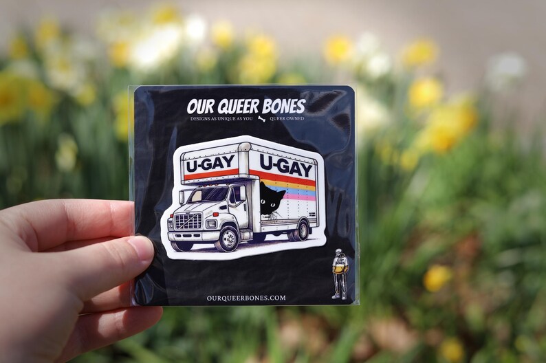 Puede incluir: Adhesivo negro con un dise&ntilde;o de cami&oacute;n blanco con el texto "U-GAY" en la parte superior y los lados. Un gato negro asoma por la ventana del cami&oacute;n. El adhesivo tambi&eacute;n incluye el texto "OUR QUEER BONES" y "DESIGNS AS UNIQUE AS YOU".