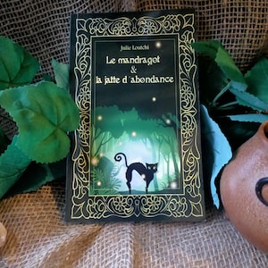 Peut inclure: Un livre noir avec une bordure dorée et un fond vert. Le titre est "Le mandragot & la jatte d'abondance" par Julie Loutchi. La couverture présente un chat noir dans un décor de forêt.