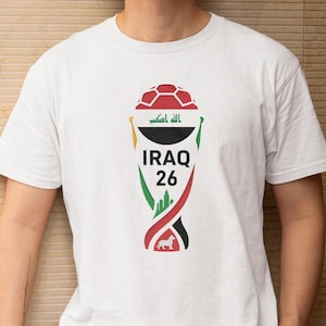 May include: White t-shirt featuring a graphic design with the text "IRAQ 26". The design incorporates elements of the Iraqi flag, a soccer ball, and a stylized trophy shape. The shirt is short-sleeved.
