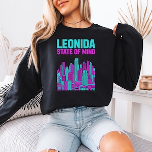 May include: A black long-sleeve sweatshirt featuring a graphic print of a city skyline in vibrant cyan and magenta. The text "LEONIDA" appears in cyan above the words "STATE OF MIND" in magenta, centered on the chest of the garment.