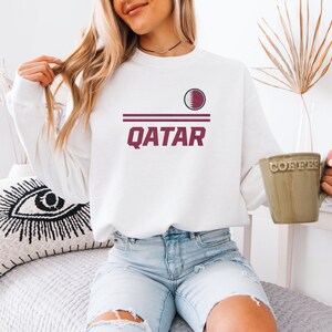 A woman wearing a white sweatshirt with the word "Qatar" printed on it, sitting on a bed and holding a coffee mug.
