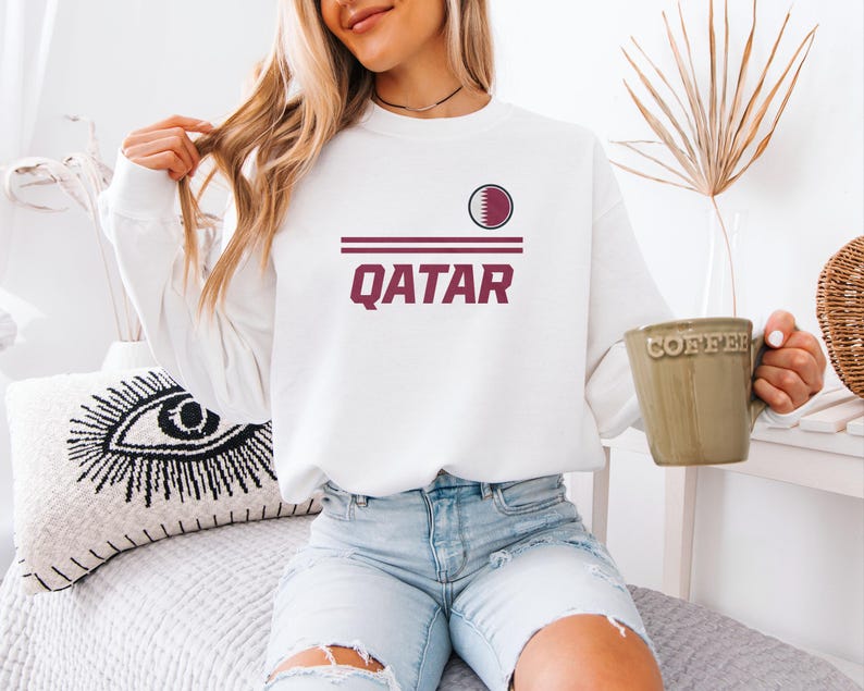 A woman wearing a white sweatshirt with the word "Qatar" printed on it, sitting on a bed and holding a coffee mug.