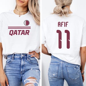A woman wearing a white t-shirt with the word "Qatar" printed on it, and a number "11" on the back. She is also wearing distressed jeans.