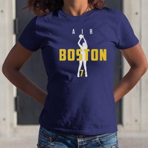 May include: Navy blue t-shirt with the word "AIR" above the word "BOSTON" in yellow block letters. A white silhouette of a basketball player shooting a ball is in the center. The number "7" is below the player.