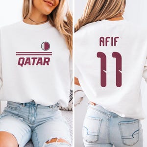 A woman wearing a white sweatshirt with the text "Qatar" and the number "11" printed on it, along with a maroon logo.