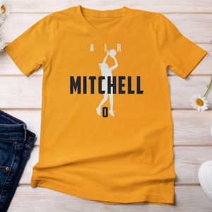 May include: Mustard yellow t-shirt featuring a white silhouette of a basketball player in action, with the name "MITCHELL" in bold black letters. Above the silhouette are the letters "A" and "R". Below the silhouette is the number "0".
