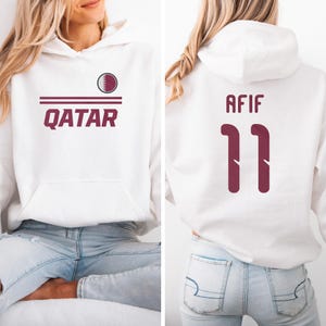 A woman wearing a white hoodie with the text "Qatar" and the number "11" printed on it.