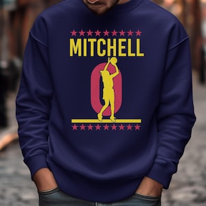 May include: Navy blue sweatshirt with the name "MITCHELL" in yellow block letters above a large maroon "0" with a yellow silhouette of a basketball player. The design is framed by rows of pink stars.