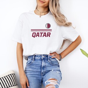A woman wearing a white t-shirt with the word "Qatar" printed on it, paired with distressed blue jeans, stands in front of a white wall.