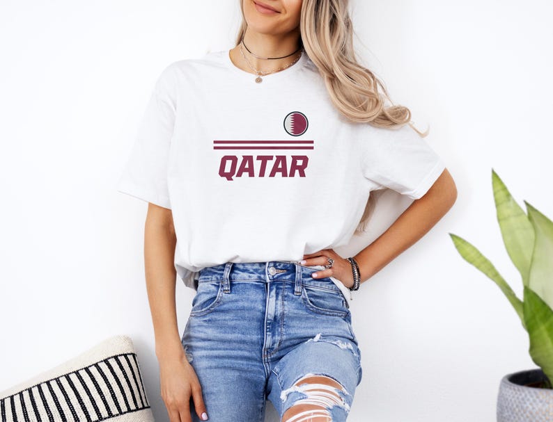 A woman wearing a white t-shirt with the word "Qatar" printed on it, paired with distressed blue jeans, stands in front of a white wall.