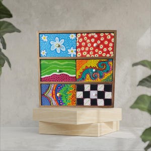 Op de afbeelding: Een kleine houten kist met zes laden, elk geschilderd met een ander kleurrijk ontwerp. De laden hebben een verscheidenheid aan patronen, waaronder bloemen, golven, spiralen en geometrische vormen.