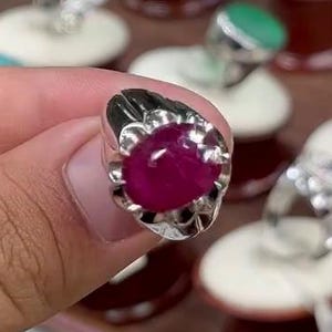 May include: A silver ring with a large, oval, deep red gemstone. The ring is held in a hand, with other rings visible in the background. The gemstone is the focal point, showcasing its rich color and setting.