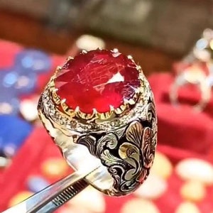 May include: A silver ring with a large, oval-shaped ruby gemstone set in a gold crown. The ring is decorated with intricate floral designs and small diamonds.