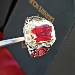 Puede incluir: Un anillo de plata con una gran piedra rectangular roja rodeada de piedras blancas más pequeñas. El anillo está engastado en una banda dorada y tiene detalles intrincados.